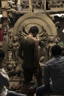 A group of people observing clay sculptures under construction in an artisanal workshop. The sculptures depict human figures and deities, including one with multiple arms. The setting has a rustic and creative atmosphere, filled with partially finished artworks and materials.
