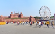 A large red sandstone fort with multiple domes and architectural features stands prominently. In front of it, a crowd of people is walking around on a spacious pavement area. To the right, a large ferris wheel with colorful cabins is visible, suggesting a festive or recreational setting. The sky is clear and the weather appears sunny.