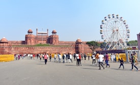 A large red sandstone fort with multiple domes and architectural features stands prominently. In front of it, a crowd of people is walking around on a spacious pavement area. To the right, a large ferris wheel with colorful cabins is visible, suggesting a festive or recreational setting. The sky is clear and the weather appears sunny.