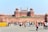 A large historical fort made of red sandstone with multiple domes and large gates. In front of the fort, many people are walking and interacting, and there are barricades marked 'Delhi Police'. The fort has an Indian flag hoisted at the top, and there is a clear blue sky in the background.