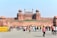 A large historical fort made of red sandstone with multiple domes and large gates. In front of the fort, many people are walking and interacting, and there are barricades marked 'Delhi Police'. The fort has an Indian flag hoisted at the top, and there is a clear blue sky in the background.