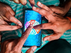 Multiple hands are pointing at a smartphone displaying a colorful board game with triangular segments, set against a teal-striped fabric background.