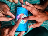 Multiple hands are pointing at a smartphone displaying a colorful board game with triangular segments, set against a teal-striped fabric background.