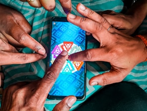 Multiple hands are pointing at a smartphone displaying a colorful board game with triangular segments, set against a teal-striped fabric background.