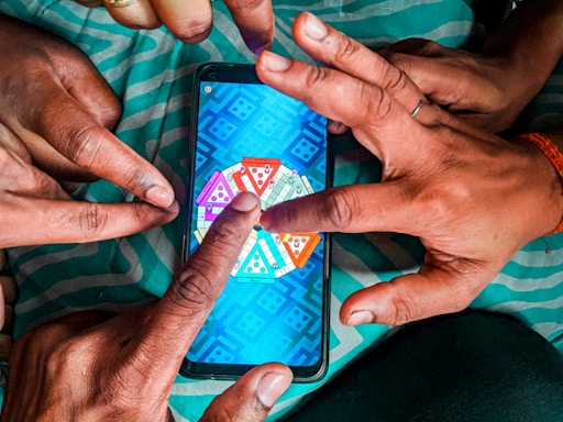 Multiple hands are pointing at a smartphone displaying a colorful board game with triangular segments, set against a teal-striped fabric background.
