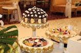 A beautifully decorated table featuring an assortment of desserts displayed on ornate stands. The central stand holds what appears to be a tower of chocolates and cream-topped pastries, surrounded by decorative gold accents. Another stand showcases a variety of colorful fruit-topped desserts, also adorned with gold embellishments. The setting is elegant, suggesting a formal event or celebration, with luxurious couches and upholstered chairs in the background.