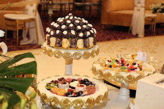A beautifully decorated table featuring an assortment of desserts displayed on ornate stands. The central stand holds what appears to be a tower of chocolates and cream-topped pastries, surrounded by decorative gold accents. Another stand showcases a variety of colorful fruit-topped desserts, also adorned with gold embellishments. The setting is elegant, suggesting a formal event or celebration, with luxurious couches and upholstered chairs in the background.