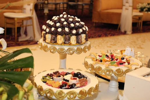 A beautifully decorated table featuring an assortment of desserts displayed on ornate stands. The central stand holds what appears to be a tower of chocolates and cream-topped pastries, surrounded by decorative gold accents. Another stand showcases a variety of colorful fruit-topped desserts, also adorned with gold embellishments. The setting is elegant, suggesting a formal event or celebration, with luxurious couches and upholstered chairs in the background.