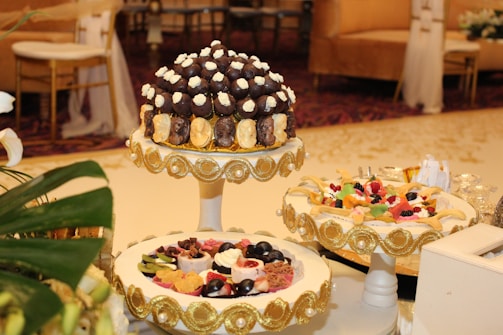 A beautifully decorated table featuring an assortment of desserts displayed on ornate stands. The central stand holds what appears to be a tower of chocolates and cream-topped pastries, surrounded by decorative gold accents. Another stand showcases a variety of colorful fruit-topped desserts, also adorned with gold embellishments. The setting is elegant, suggesting a formal event or celebration, with luxurious couches and upholstered chairs in the background.