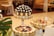 A beautifully decorated table featuring an assortment of desserts displayed on ornate stands. The central stand holds what appears to be a tower of chocolates and cream-topped pastries, surrounded by decorative gold accents. Another stand showcases a variety of colorful fruit-topped desserts, also adorned with gold embellishments. The setting is elegant, suggesting a formal event or celebration, with luxurious couches and upholstered chairs in the background.