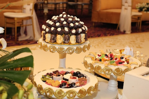 A beautifully decorated table featuring an assortment of desserts displayed on ornate stands. The central stand holds what appears to be a tower of chocolates and cream-topped pastries, surrounded by decorative gold accents. Another stand showcases a variety of colorful fruit-topped desserts, also adorned with gold embellishments. The setting is elegant, suggesting a formal event or celebration, with luxurious couches and upholstered chairs in the background.