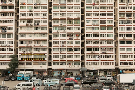 An urban residential building filled with rows of windows, air conditioning units, and laundry hanging outside. Vehicles are parked in front of the building, including cars and vans, suggesting a bustling residential area. The building is marked with signs in different languages, likely indicating businesses or services located on the ground floor.