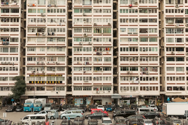 An urban residential building filled with rows of windows, air conditioning units, and laundry hanging outside. Vehicles are parked in front of the building, including cars and vans, suggesting a bustling residential area. The building is marked with signs in different languages, likely indicating businesses or services located on the ground floor.