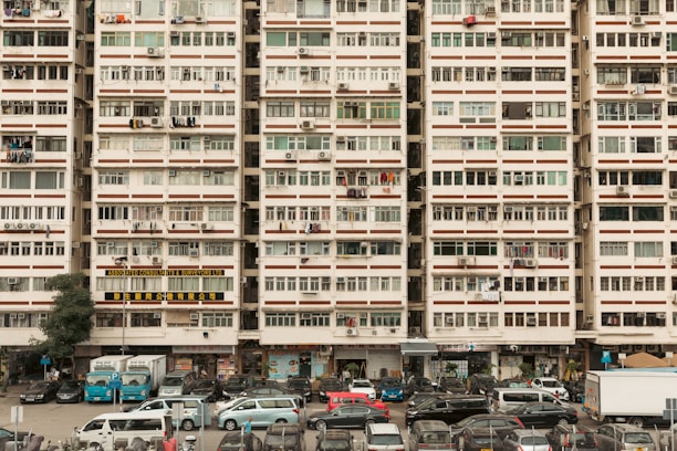 An urban residential building filled with rows of windows, air conditioning units, and laundry hanging outside. Vehicles are parked in front of the building, including cars and vans, suggesting a bustling residential area. The building is marked with signs in different languages, likely indicating businesses or services located on the ground floor.