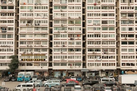 An urban residential building filled with rows of windows, air conditioning units, and laundry hanging outside. Vehicles are parked in front of the building, including cars and vans, suggesting a bustling residential area. The building is marked with signs in different languages, likely indicating businesses or services located on the ground floor.