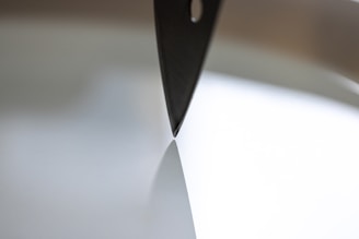 A close-up shot of a knife blade positioned vertically against a flat, reflective surface, creating a sharp contrast between the metallic edge and the smooth background.