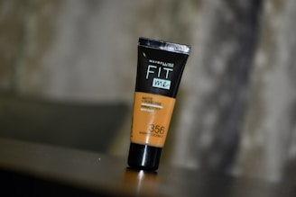 A tube of Maybelline Fit Me Matte + Poreless foundation is placed on a reflective surface with a blurred background. The tube is upright, featuring a warm coconut shade labeled as number 356.