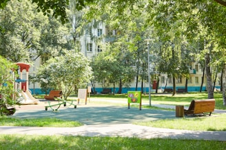 A sunny park scene with a playground featuring a slide and seesaw surrounded by lush green trees and grass. In the background, there are apartment buildings with balconies, partially obscured by the foliage. The area is well-lit, with benches and pathways providing a peaceful and inviting atmosphere.