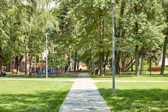 A sunny park scene with a walkway leading through lush green grass and surrounded by tall trees. A playground with colorful equipment is partially visible among the trees. Lampposts line the edges of the path, and there is a red brick building partially obscured in the background.