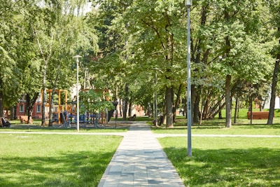 A sunny park scene with a walkway leading through lush green grass and surrounded by tall trees. A playground with colorful equipment is partially visible among the trees. Lampposts line the edges of the path, and there is a red brick building partially obscured in the background.