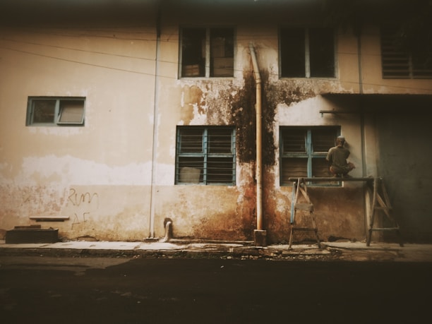 An old, weathered building facade with peeling paint and mold streaks, featuring blue-framed windows. A person sits on a makeshift platform or scaffolding, appearing to work on one of the windows.