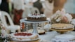 A variety of cakes are displayed elegantly on a table, with one chocolate cake prominently placed on a decorative stand adorned with crystal-like ornaments. Floral arrangements with pink and white flowers are in the background, adding a touch of elegance to the setting. The cakes have various toppings, including whipped cream and fruit.