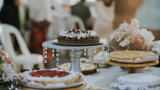 A variety of cakes are displayed elegantly on a table, with one chocolate cake prominently placed on a decorative stand adorned with crystal-like ornaments. Floral arrangements with pink and white flowers are in the background, adding a touch of elegance to the setting. The cakes have various toppings, including whipped cream and fruit.