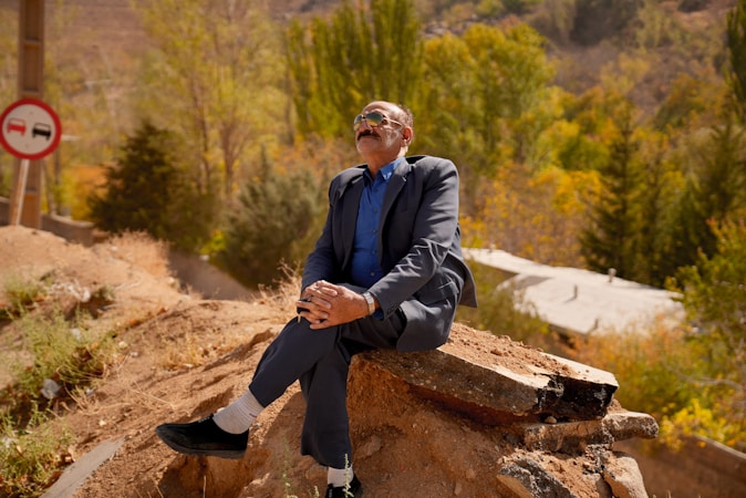 An older man in a suit and tie is sitting on a rock or concrete slab. He is wearing sunglasses and appears to be relaxed with his hands clasped on his lap. The background features a mix of greenery and autumn-colored trees, with a road sign visible on the left side of the image.