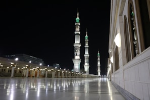 A night-time scene featuring an illuminated mosque courtyard with multiple tall minarets adorned with intricate designs. The structure on the right shows arched windows and decorative lighting. The smooth, reflective flooring enhances the serene atmosphere.