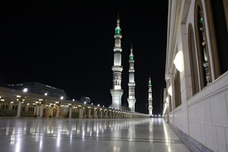 A night-time scene featuring an illuminated mosque courtyard with multiple tall minarets adorned with intricate designs. The structure on the right shows arched windows and decorative lighting. The smooth, reflective flooring enhances the serene atmosphere.