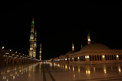 A nighttime scene featuring an illuminated mosque with multiple minarets adorned with green and white lights. The courtyard is vast and well-lit, with reflections visible on the polished marble surface. The architecture displays large domes and arches, emphasizing the grandeur and serenity of the setting.