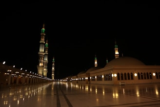 A nighttime scene featuring an illuminated mosque with multiple minarets adorned with green and white lights. The courtyard is vast and well-lit, with reflections visible on the polished marble surface. The architecture displays large domes and arches, emphasizing the grandeur and serenity of the setting.