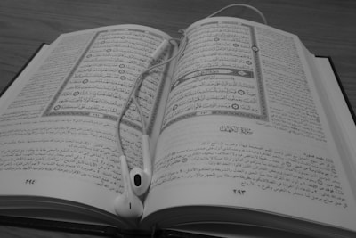 A black and white image showing an open book with Arabic text on both pages. Earphones are placed on top of the book, with the wires casually laid out.