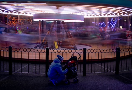 A blurred carousel spins in a circular motion, surrounded by festive lights and decorations. In the foreground, a person dressed in a blue winter coat is kneeling beside a stroller and appears to be adjusting or interacting with something inside. The scene is bathed in vibrant, multi-colored lights, creating an energetic and whimsical atmosphere.