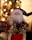 A decorative figure of Santa Claus is positioned in front of a blurred, brightly lit Christmas tree. The Santa figure is adorned with red, white, and gold clothing, featuring intricate patterns and embellishments. The background of the image is filled with colorful bokeh lights, adding to the festive atmosphere.