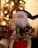 A decorative figure of Santa Claus is positioned in front of a blurred, brightly lit Christmas tree. The Santa figure is adorned with red, white, and gold clothing, featuring intricate patterns and embellishments. The background of the image is filled with colorful bokeh lights, adding to the festive atmosphere.
