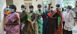 A group of elderly individuals wearing face masks stand in a line indoors. They are dressed in traditional clothing, holding documents or bags, indicating they might be waiting for a service or appointment. The room has neutral walls and some natural light coming in.