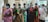 A group of elderly individuals wearing face masks stand in a line indoors. They are dressed in traditional clothing, holding documents or bags, indicating they might be waiting for a service or appointment. The room has neutral walls and some natural light coming in.