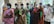 A group of elderly individuals wearing face masks stand in a line indoors. They are dressed in traditional clothing, holding documents or bags, indicating they might be waiting for a service or appointment. The room has neutral walls and some natural light coming in.