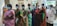 A group of elderly individuals wearing face masks stand in a line indoors. They are dressed in traditional clothing, holding documents or bags, indicating they might be waiting for a service or appointment. The room has neutral walls and some natural light coming in.