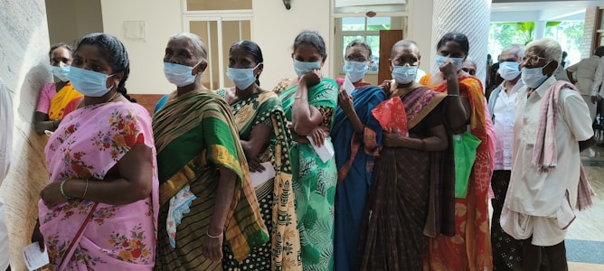 A group of elderly individuals wearing face masks stand in a line indoors. They are dressed in traditional clothing, holding documents or bags, indicating they might be waiting for a service or appointment. The room has neutral walls and some natural light coming in.