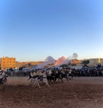 A group of riders on horseback are performing a traditional charge, with one rider firing a rifle that releases smoke and flames. Spectators are gathered around the dusty arena, and buildings are visible in the background under the clear blue sky.