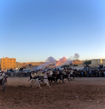 A group of riders on horseback are performing a traditional charge, with one rider firing a rifle that releases smoke and flames. Spectators are gathered around the dusty arena, and buildings are visible in the background under the clear blue sky.