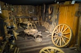 The room is filled with antique wooden tools and wagon wheels, displayed against wooden walls and floors. Various woodworking tools hang on the walls, creating a rustic and historical atmosphere similar to an old blacksmith or craftsman shop.