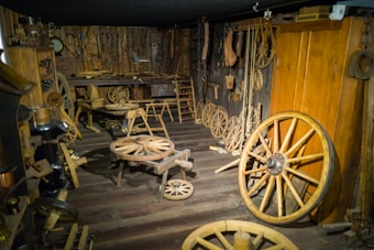 The room is filled with antique wooden tools and wagon wheels, displayed against wooden walls and floors. Various woodworking tools hang on the walls, creating a rustic and historical atmosphere similar to an old blacksmith or craftsman shop.