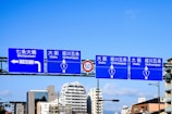 Blue road signs with white text and directional arrows are displayed above a street, indicating directions to various locations in Osaka. The background features a clear blue sky and a cityscape with modern buildings.