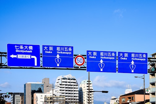 Blue road signs with white text and directional arrows are displayed above a street, indicating directions to various locations in Osaka. The background features a clear blue sky and a cityscape with modern buildings.
