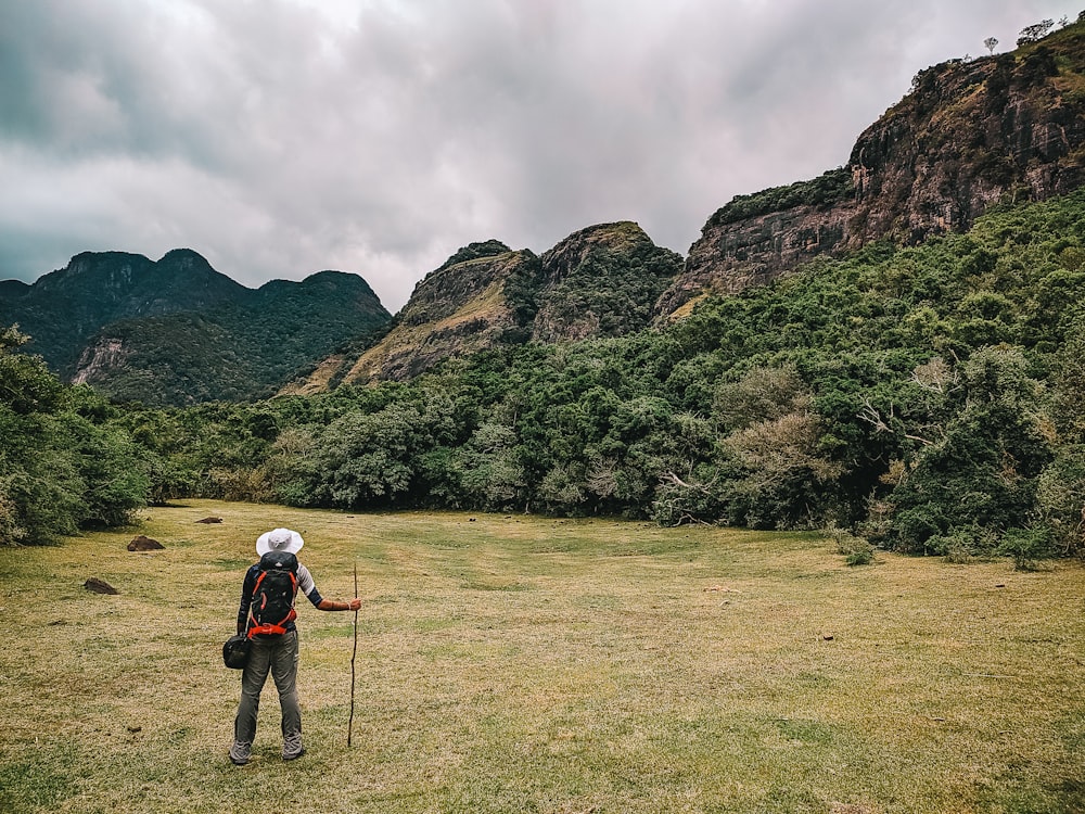 a man with a backpack standing in a field with trees and mountains in the background