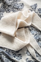 A soft, knitted cream-colored scarf arranged elegantly on a patterned fabric background. The fabric features intricate floral designs in black or dark blue, creating a striking contrast with the scarf.