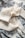 A soft, knitted cream-colored scarf arranged elegantly on a patterned fabric background. The fabric features intricate floral designs in black or dark blue, creating a striking contrast with the scarf.